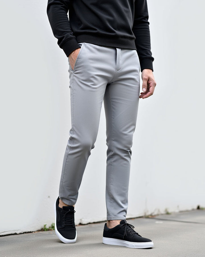 Lysegrå chinos båret med sort sweatshirt og sorte sneakers foran en hvid væg, set forfra og let fra siden. 2