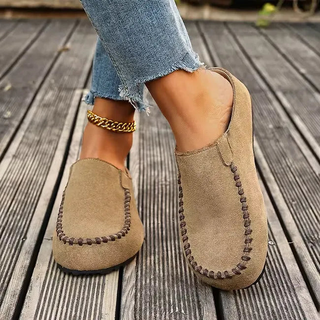 Lysebrune slip-ons båret på træterrasse, set skråt forfra, så den lukkede tå, syningen og den flade sål ses. 5