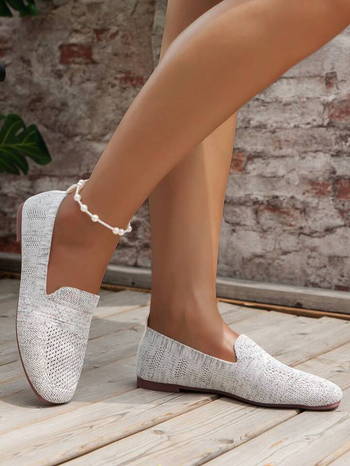 Lysegrå strikkede slip-on sko på model med perleankelkæde, set fra siden foran murstensvæg. 14