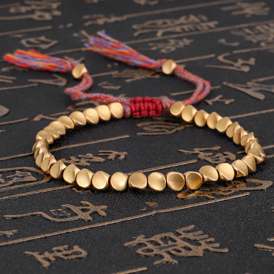 Goldenes Armband mit runden Perlen und bunten Quasten auf schwarzem Hintergrund mit Schriftzeichen.
