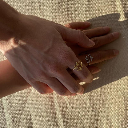 Zwei Hände mit goldenen und silbernen Blatt-Ringen auf beigem Stoff.