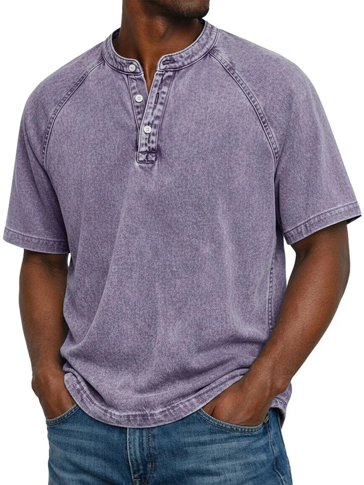 Mand i lilla vasket Henley-T-shirt med raglanærmer og knaplukning ved halsen, hænderne i lommerne på blå jeans. 1