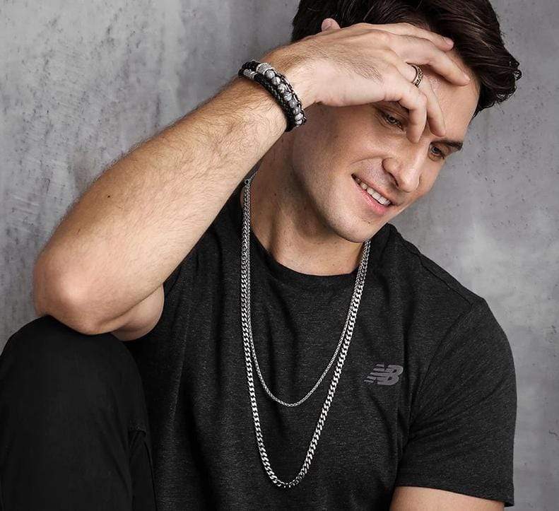 Mann mit schwarzem T-Shirt trägt silberne Kette und Steinperlenarmband, lächelt, sitzt entspannt.