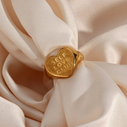 Goldener Herzring mit Gravur "Self Love Club" auf beigem Stoff, Schmuck für Selbstliebe.