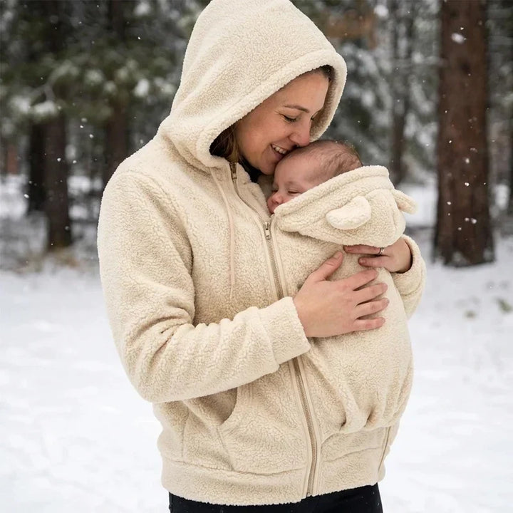 Kvinde i cremefarvet sherpa-trøje med hætte kysser sin baby i bærepanelet, mens det sner i skoven. 8