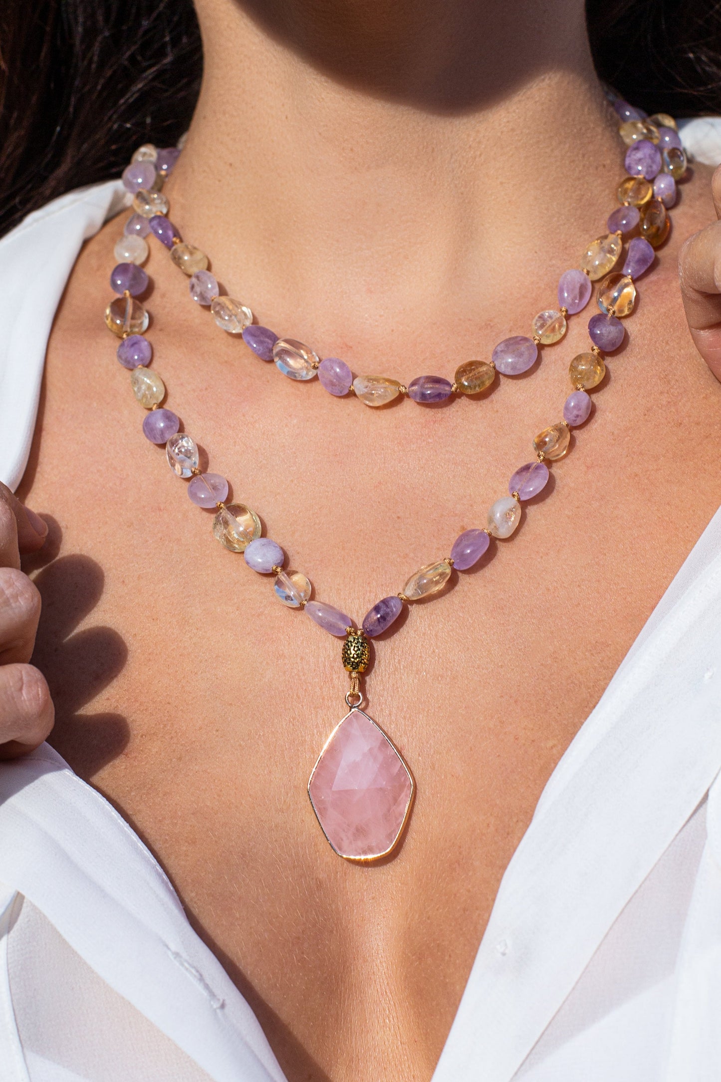 Frau mit zweireihiger Halskette aus lila und goldenen Edelsteinen, rosa Anhänger, weißes Hemd.