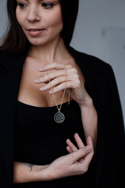 Frau mit schwarzem Blazer hält goldene Halskette mit rundem Anhänger.