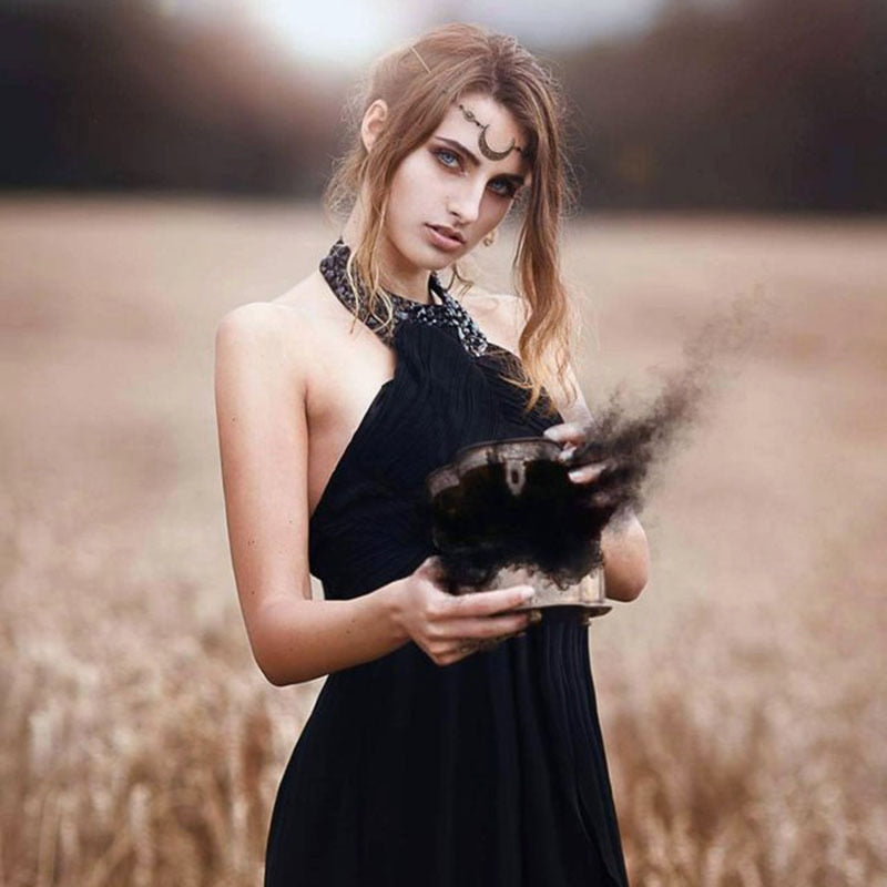 Frau in schwarzem Kleid hält rauchende Schatulle auf Feld, mystische Atmosphäre.