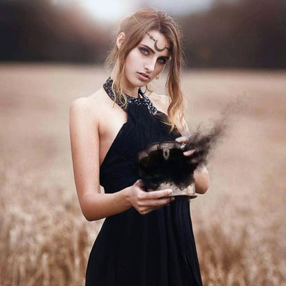 Frau in schwarzem Kleid hält rauchende Schatulle auf Feld, mystische Atmosphäre.