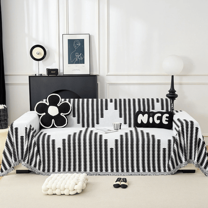 Sofa dækket af sort-hvidt chenille-betræk med lodret bladmønster og frynser, set forfra i stue med blomster- og skriftpuder. 1