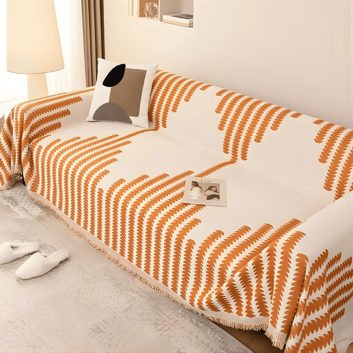 Sofabetræk i orange og creme med bladmønster og frynser, set fra siden på en sofa med mønstret pude. 4