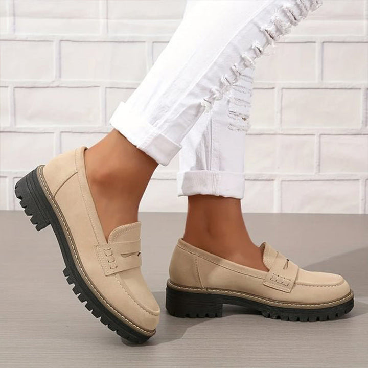 Beige loafers med kraftig sort sål set fra siden, båret med hvide oprullede jeans foran hvid murstensvæg. 3