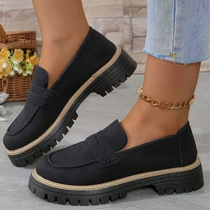 Sorte loafers med kraftig platformsål og beige kant, båret med guldfarvet ankelkæde ved siden af kurv med blomster. 12